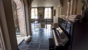 A spacious room with a piano, a dining area with a table and chairs, and a large window with a view of a brick wall. - B&B Filemon & Baucis (Bruges)