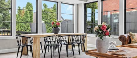Une pièce spacieuse avec une grande table à manger, plusieurs chaises et un canapé. De grandes fenêtres offrent une vue sur les arbres et un mur en briques.