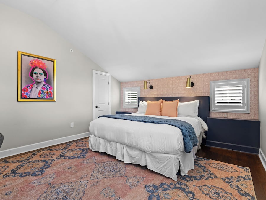 A bedroom with a bed, a patterned rug, a wall-mounted light fixture, and a door.