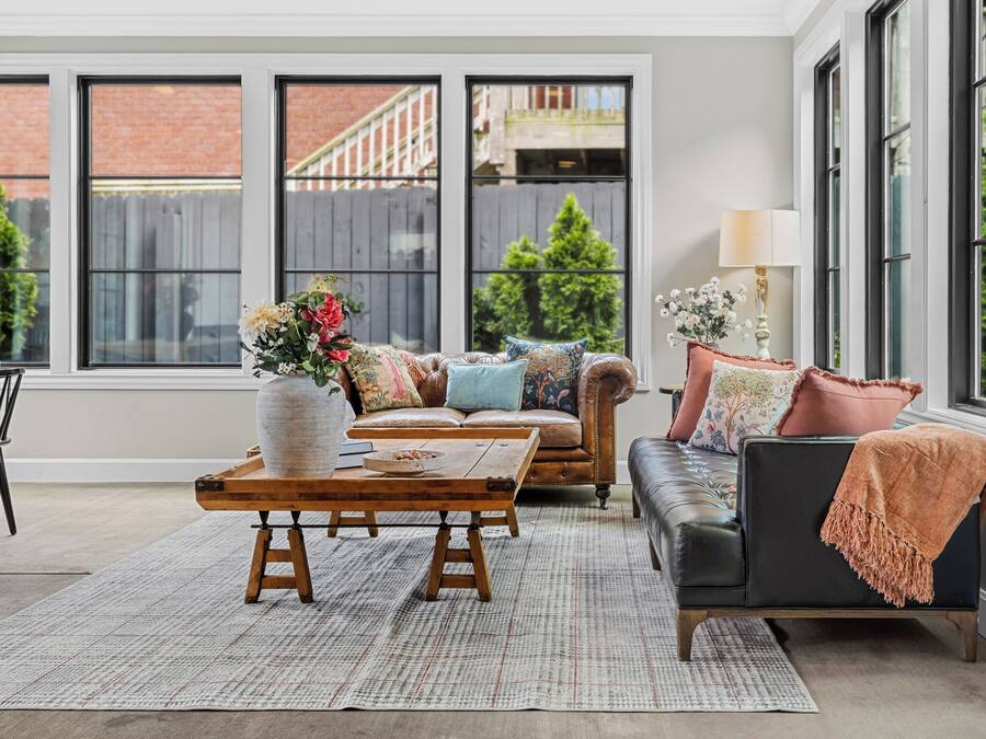 Una sala de estar acogedora con un sofá, una mesa de centro y una mesa de comedor. Hay ventanas que dan a una pared de ladrillo y follaje al exterior.