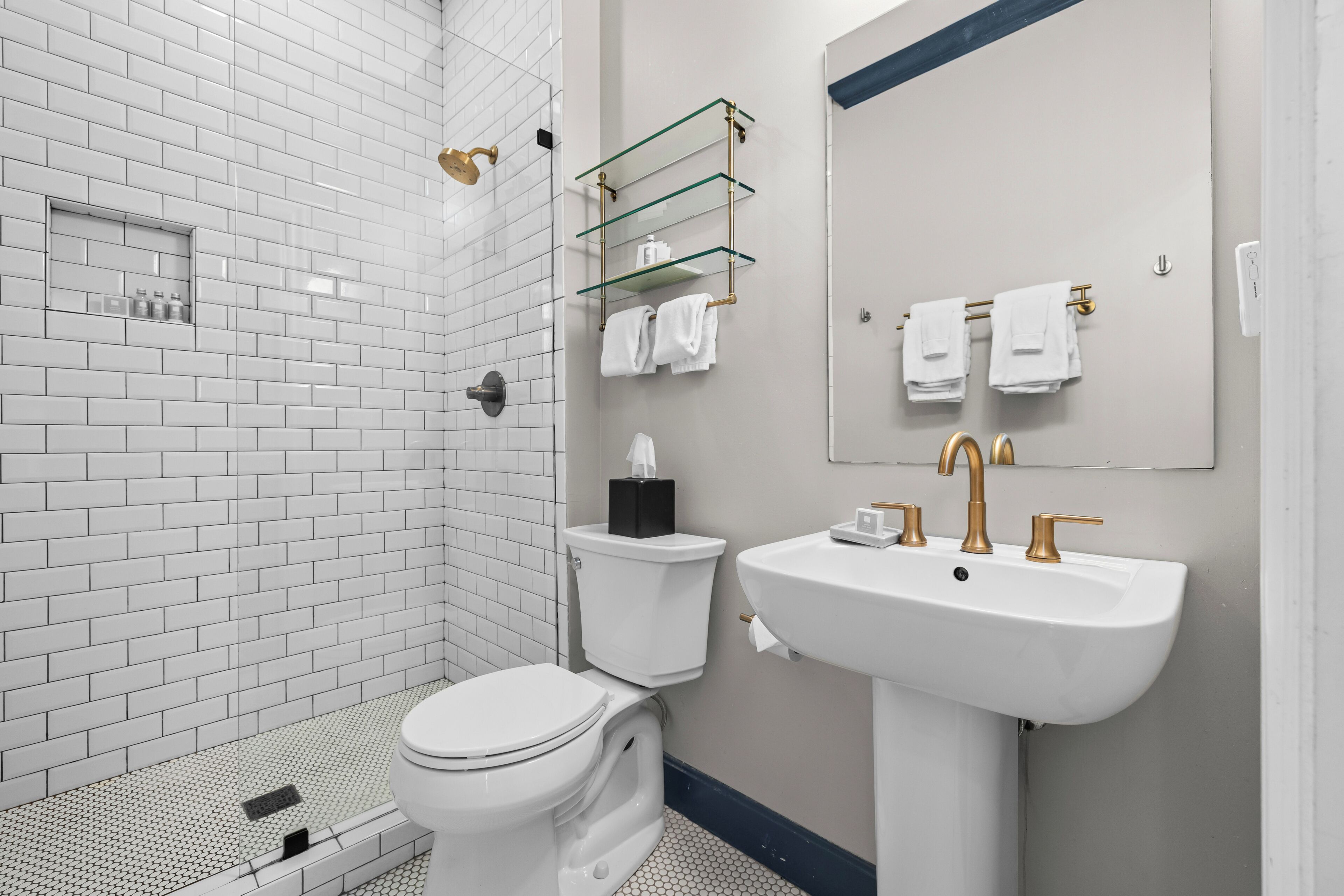 Un baño moderno con ducha de azulejos blancos, inodoro, lavamanos con detalles dorados y repisa de vidrio con artículos de aseo.