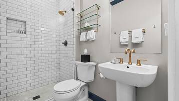 Un baño moderno con ducha de azulejos blancos, inodoro, lavamanos con detalles dorados y repisa de vidrio con artículos de aseo.