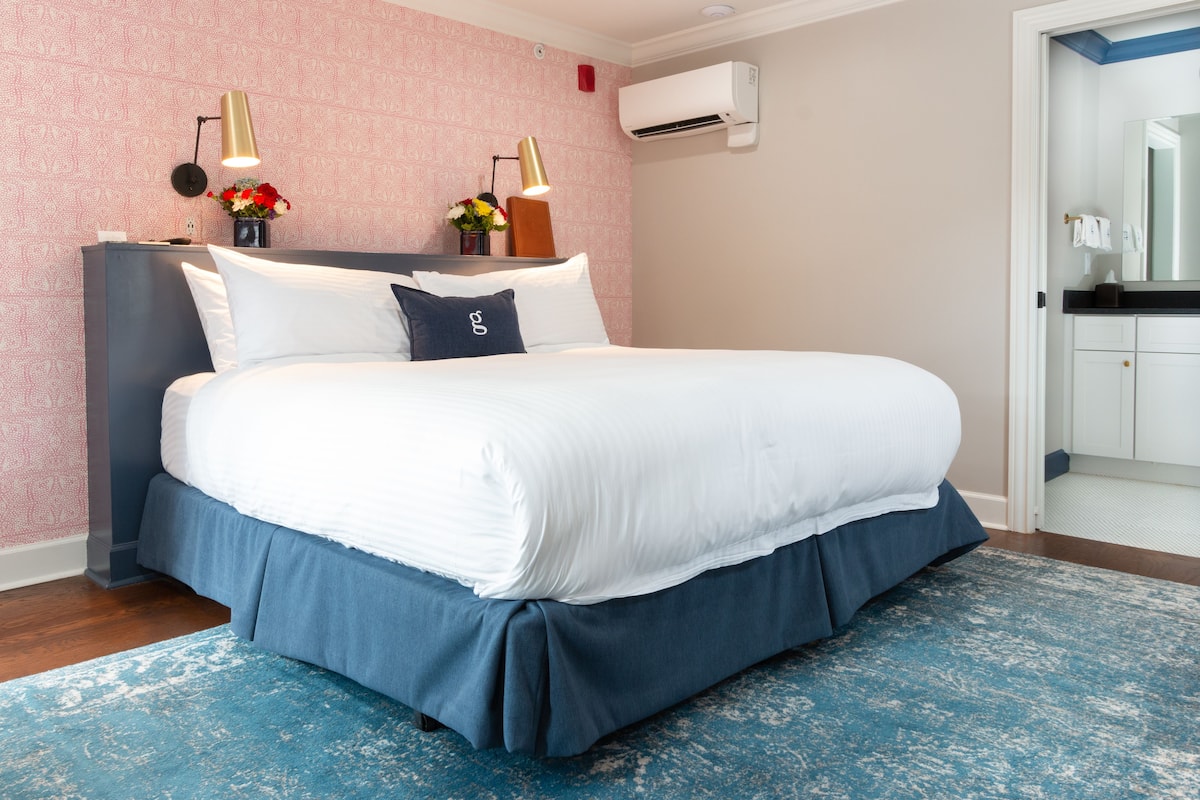 A neatly made bed with white and blue bedding, a blue accent rug, and two wall-mounted lamps.
