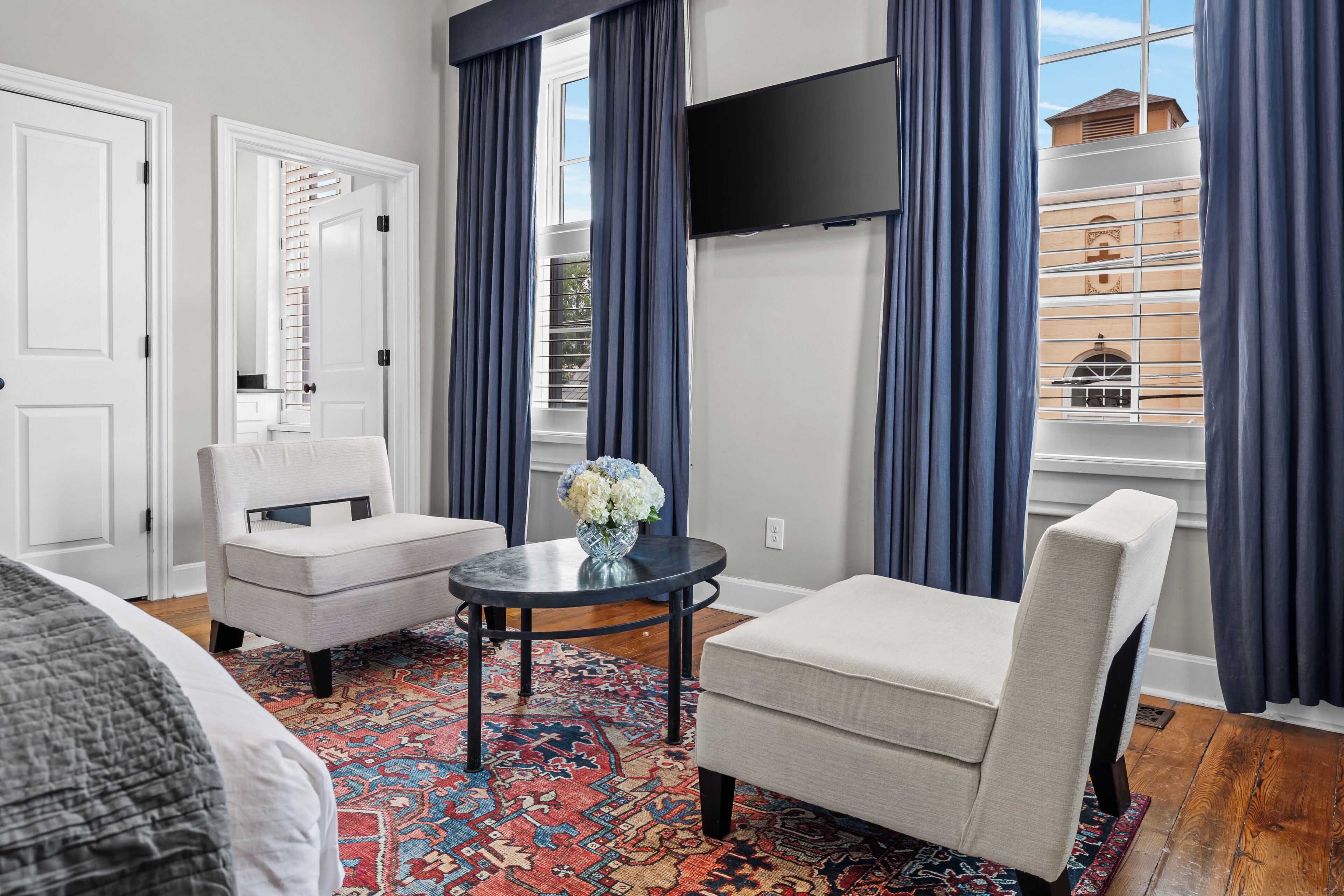 Ein Hotelzimmer mit einem Bett, zwei Sesseln, einem kleinen Tisch mit einer Vase und Blick auf die Stadt durch große Fenster.