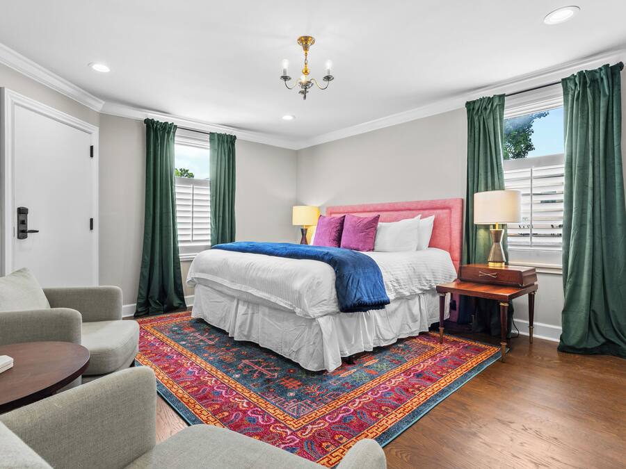 Un dormitorio con cama, mesita de noche, una silla, alfombra y ventana con cortinas.