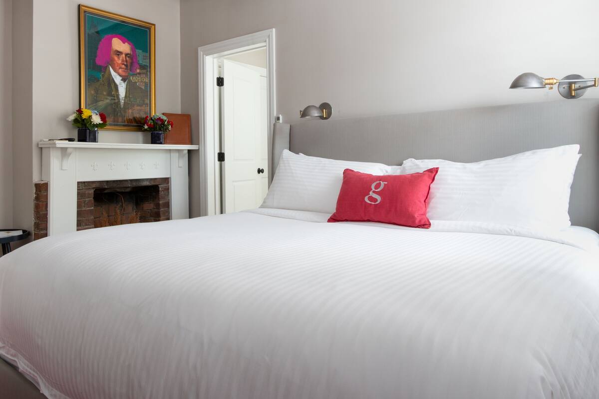 A neatly arranged bedroom with a large bed, a fireplace, a portrait, and a nightstand.