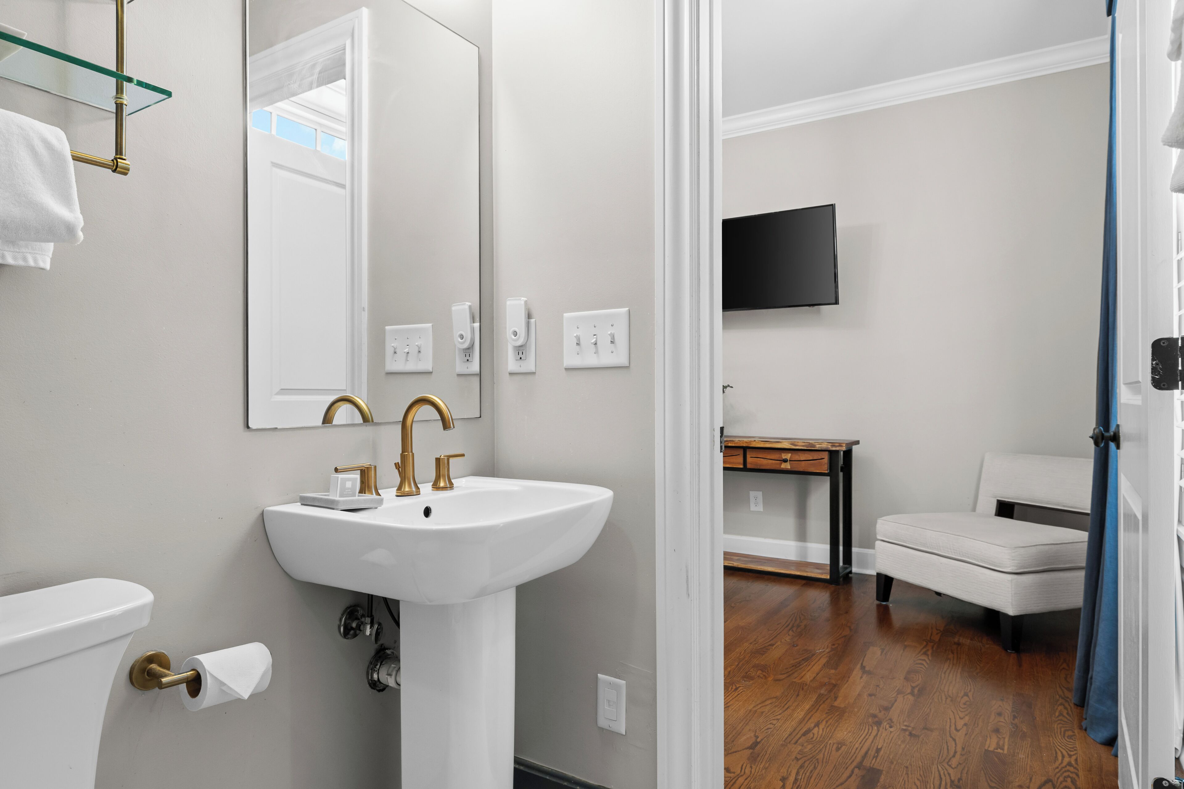 A modern bathroom with a white sink, gold faucets, and a large mirror. There is a toilet with a paper towel holder and a towel hanging on the wall. A television is mounted on the wall, and a wooden bench is visible in the background.