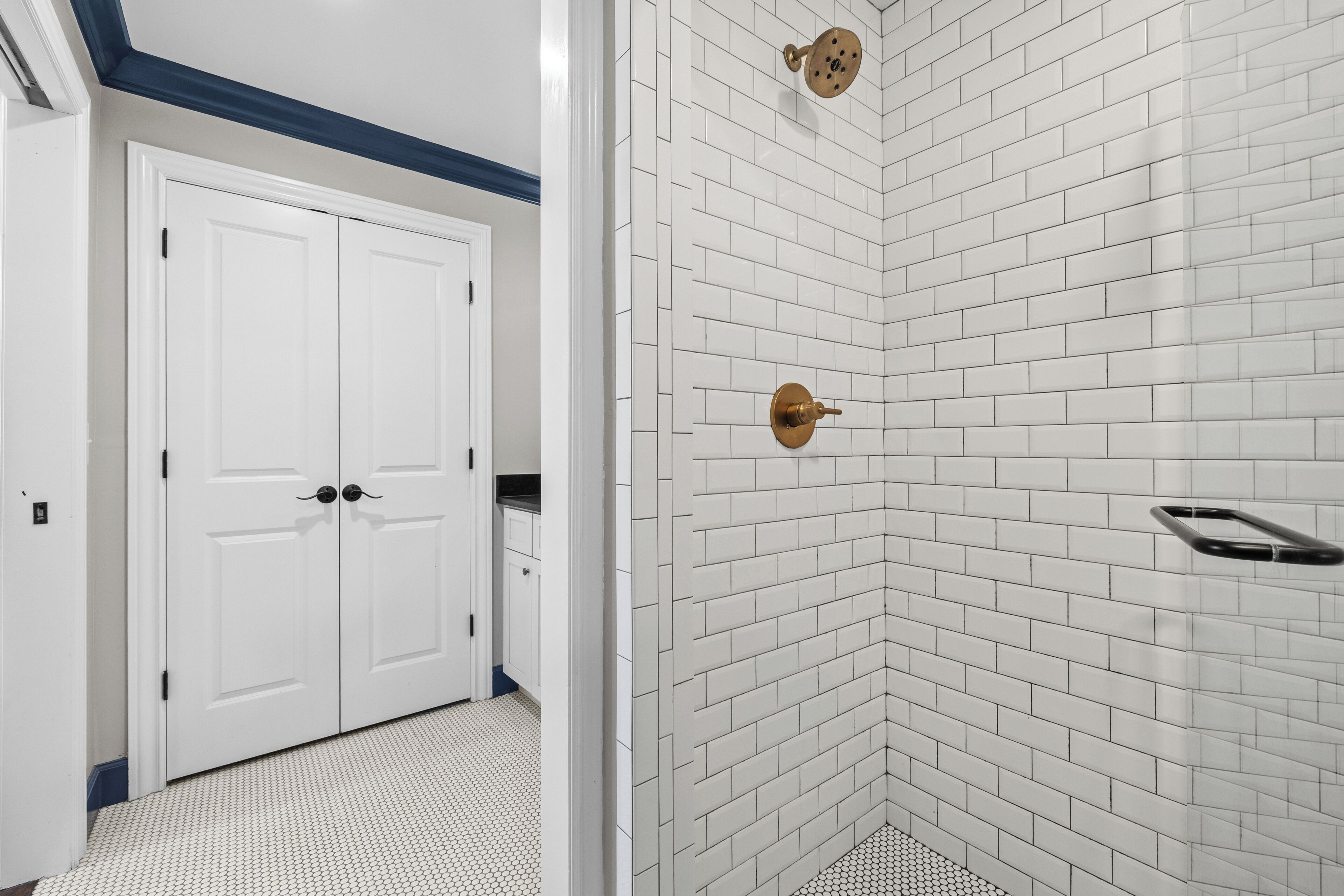 A bathroom with white subway tiles, a shower with a gold showerhead, and a white door with a black handle.