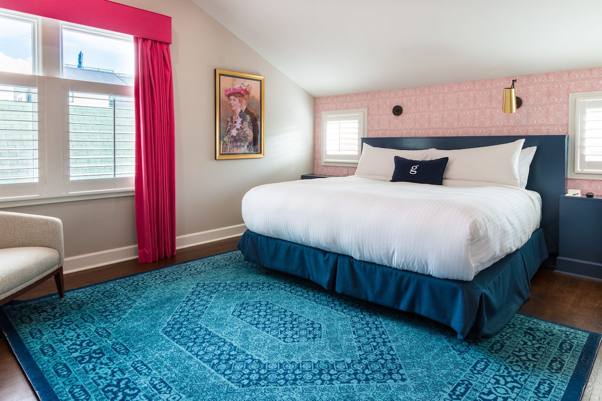 A bedroom with a large bed, a blue patterned rug, a pink wall, and a window with blinds.