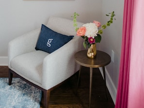 A room with a white armchair, a small round table with a vase of flowers, and a blue cushion with a logo.