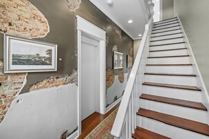 Una escalera con escalones de madera y barandillas blancas que conduce a una habitación con pared de ladrillo y un cuadro enmarcado.