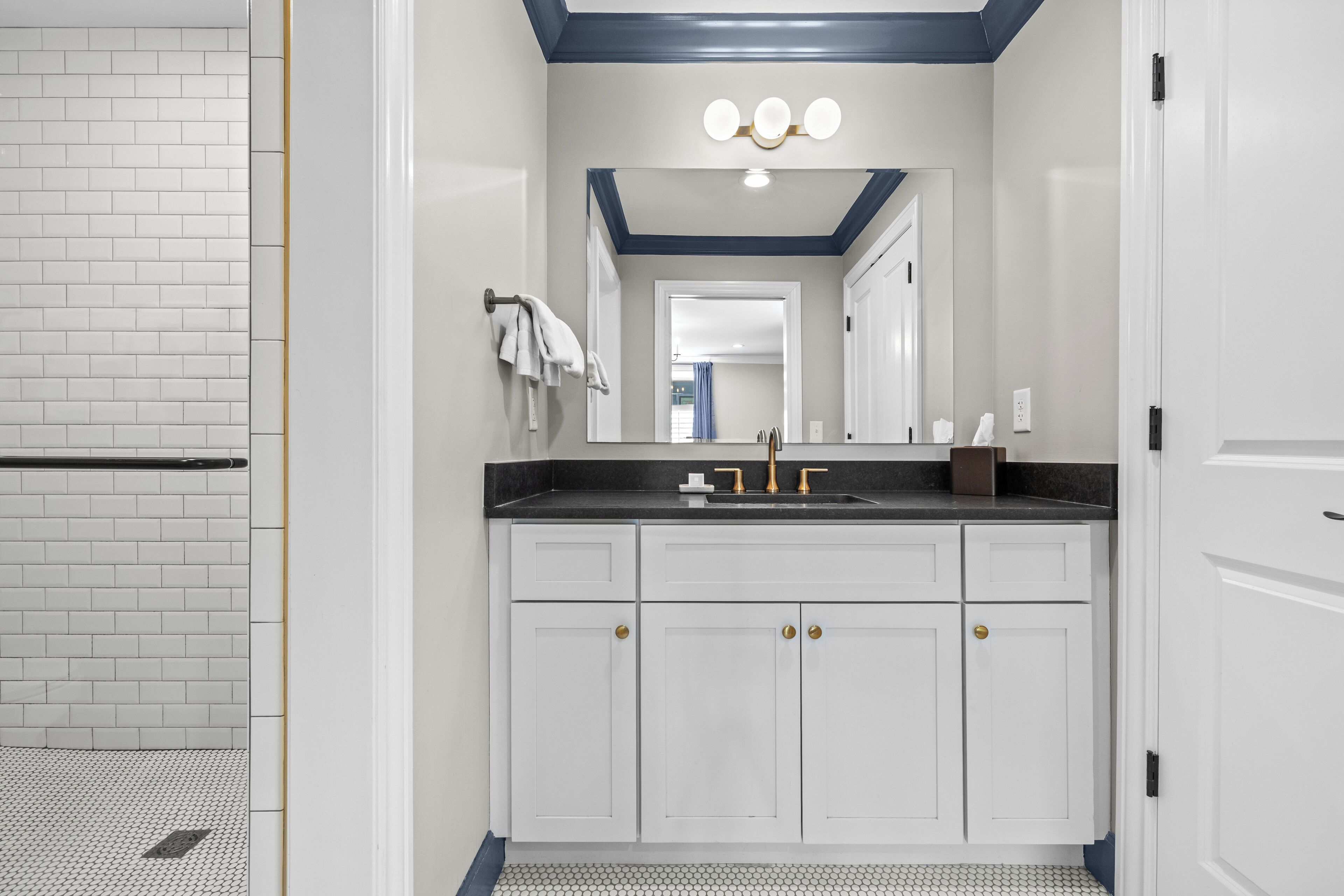A bathroom with a white tiled wall, a black countertop, a sink with gold fixtures, and a mirror above the sink.