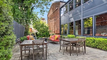 Un patio al aire libre con mesas y sillas de madera, un muro de ladrillo y un edificio moderno con amplios ventanales.