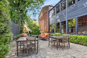An outdoor patio with wooden tables and chairs, a brick wall, and a modern building with large windows.