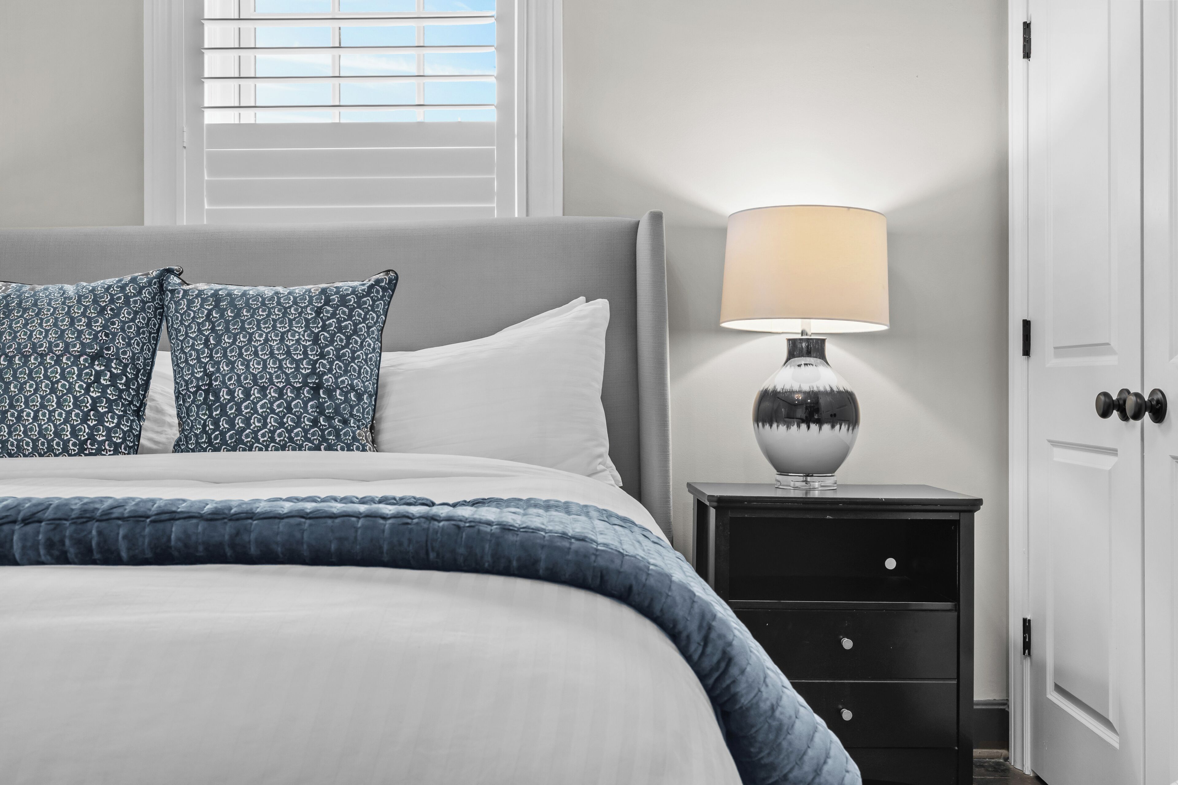Una cama bien hecha con un cojín de diseño azul y blanco, una lámpara de noche y una mesita de noche con cajones.
