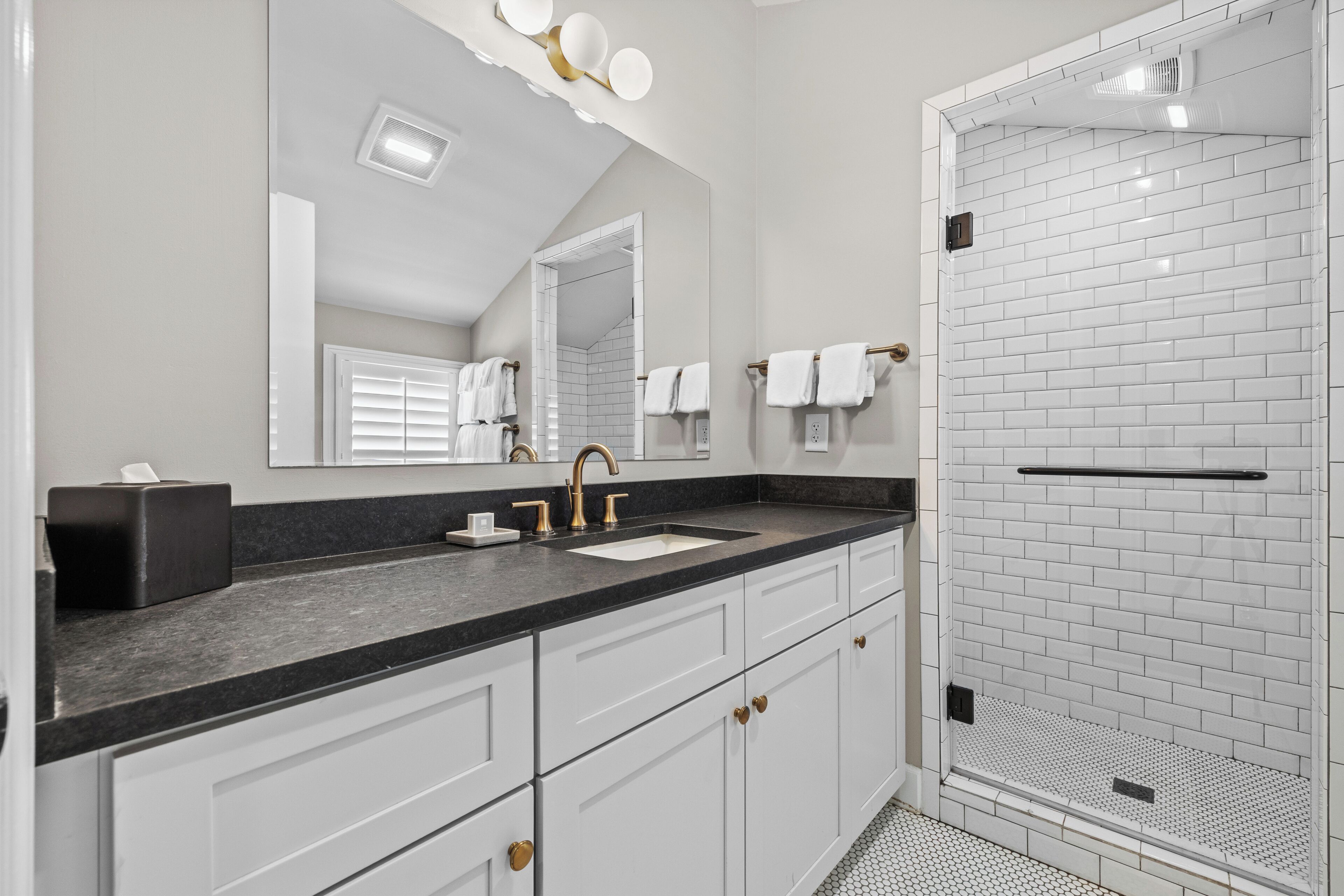 A modern bathroom with a black countertop, white cabinets, a large mirror, and a shower with white tiles.