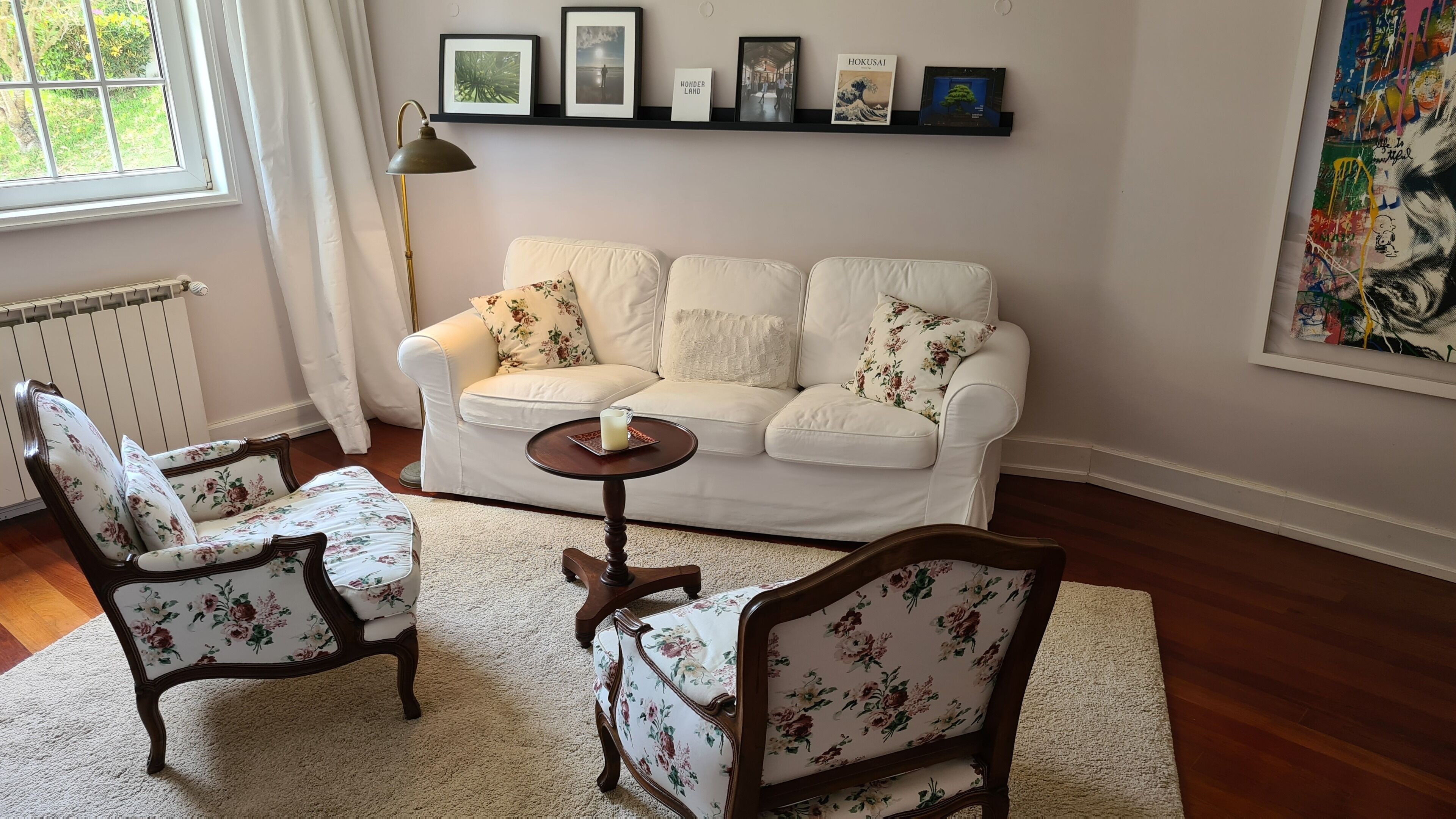 Een woonkamer met een witte bank, twee armstoelen met bloemenprint, een klein rond tafeltje met een kaars, en een radiator.