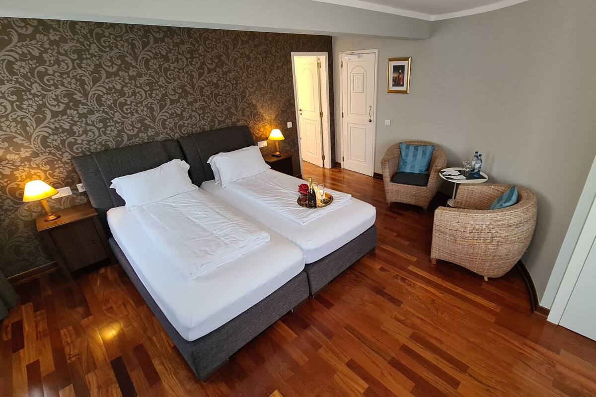 Habitació d'hotel amb un llit doble, dues taules de nit amb làmpades, una taula rodona petita amb una cadira i una porta que dona a una altra habitació.