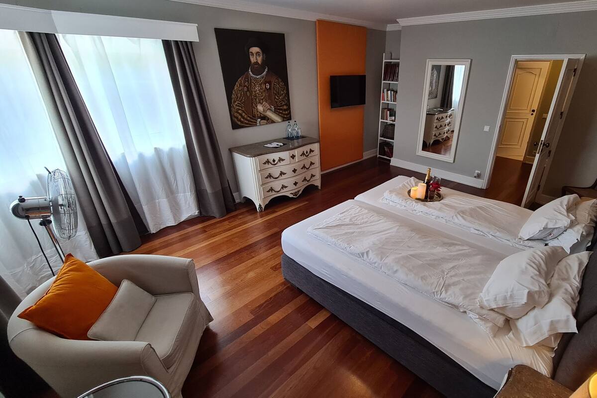 Habitació d'hotel amb un llit gran, una taula de nit, una cadira, un armari, un mirall i una finestra amb cortines.