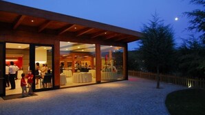 Espai per a esdeveniments a l'aire lliure amb zona coberta i parets de vidre, envoltat d'arbres i un camí de grava.