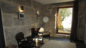 Habitació amb parets de pedra, dues cadires, una taula petita amb un vas, una cistella i vista a zones verds a través d'una finestra.