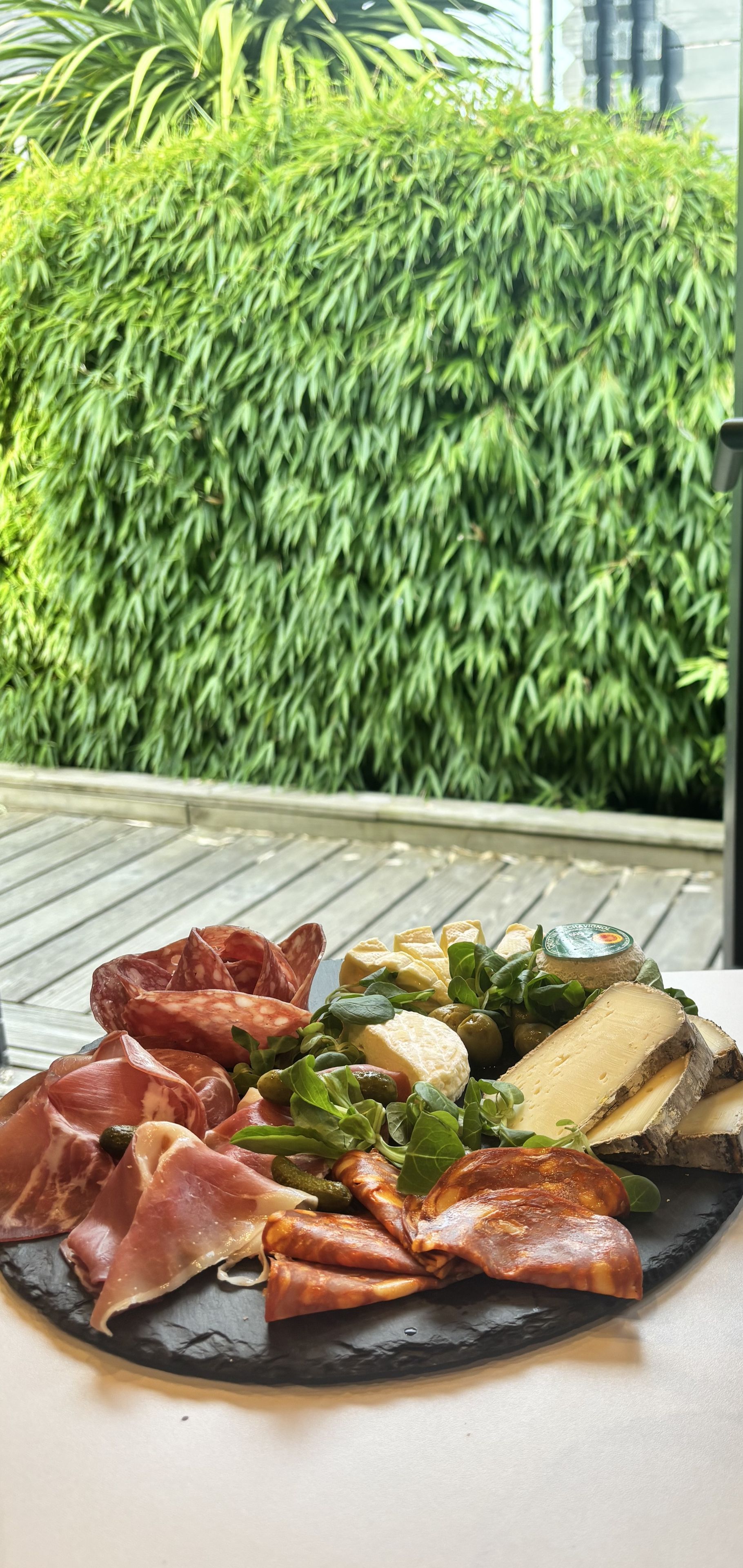 Een charcuteriebord met verschillende vleessoorten en kazen op een leisteenbord, buiten gezet met een groene haag op de achtergrond.