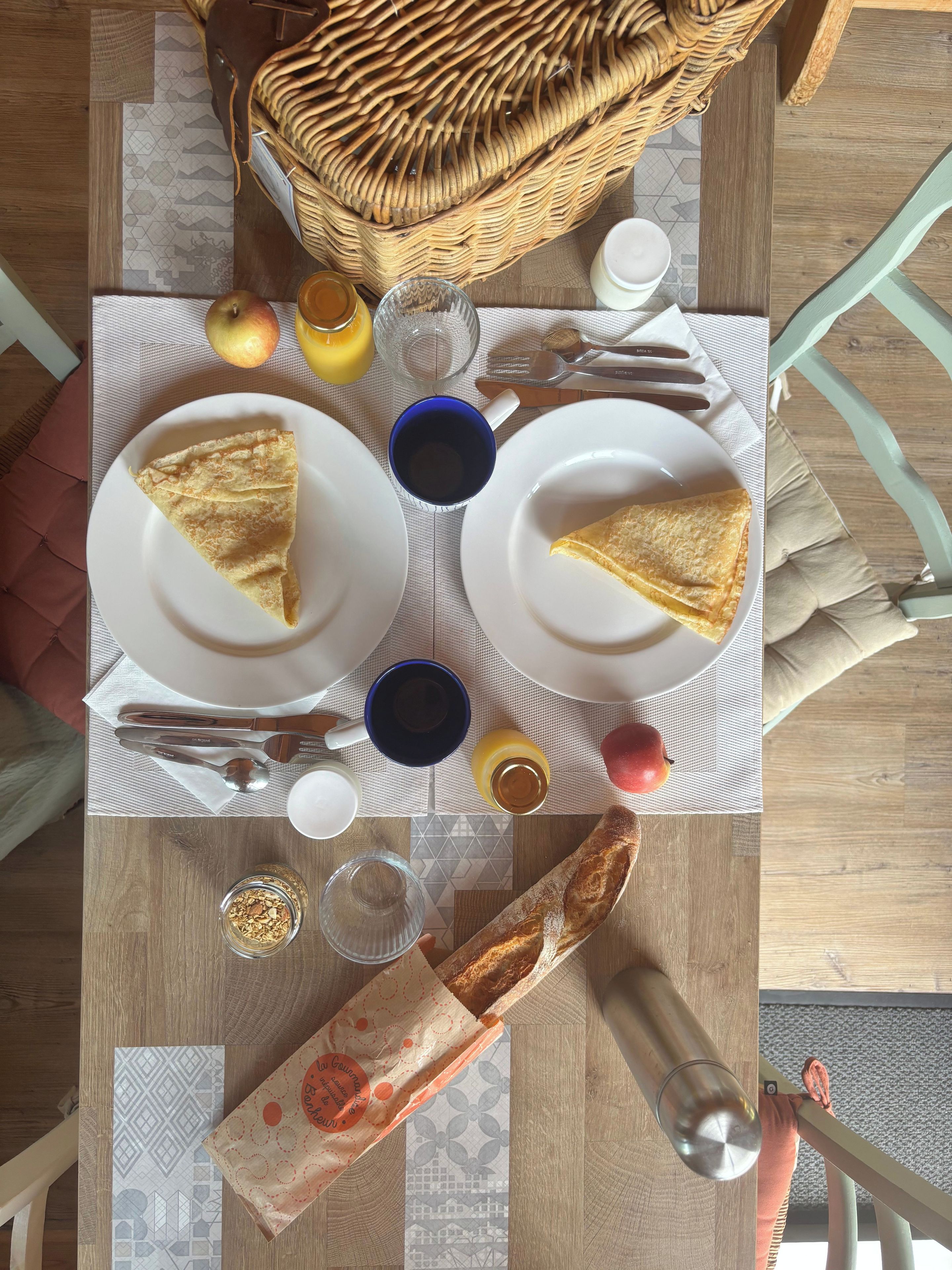 A breakfast setting with a wicker basket, plates with crepes, a baguette, and various breakfast items on a table.