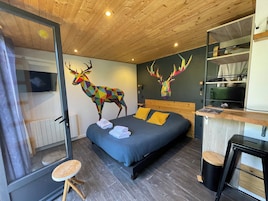 Un dormitorio moderno con techo de madera, una cama con ropa de cama azul, dos almohadas amarillas, un cuadro de un ciervo colorido en la pared y un taburete de madera pequeño.