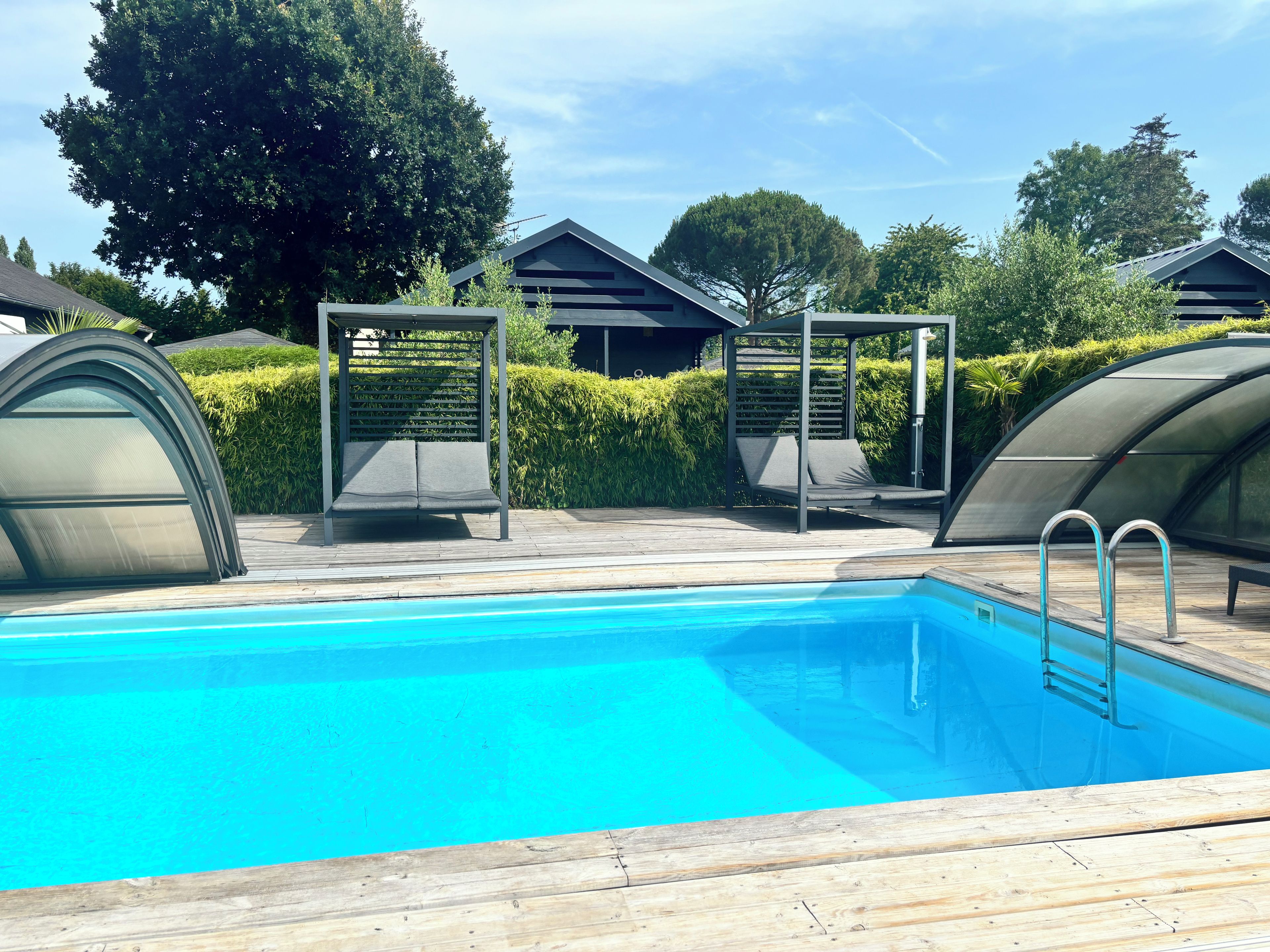 Una piscina con cubierta de madera, dos zonas de estar cubiertas y un seto al fondo.
