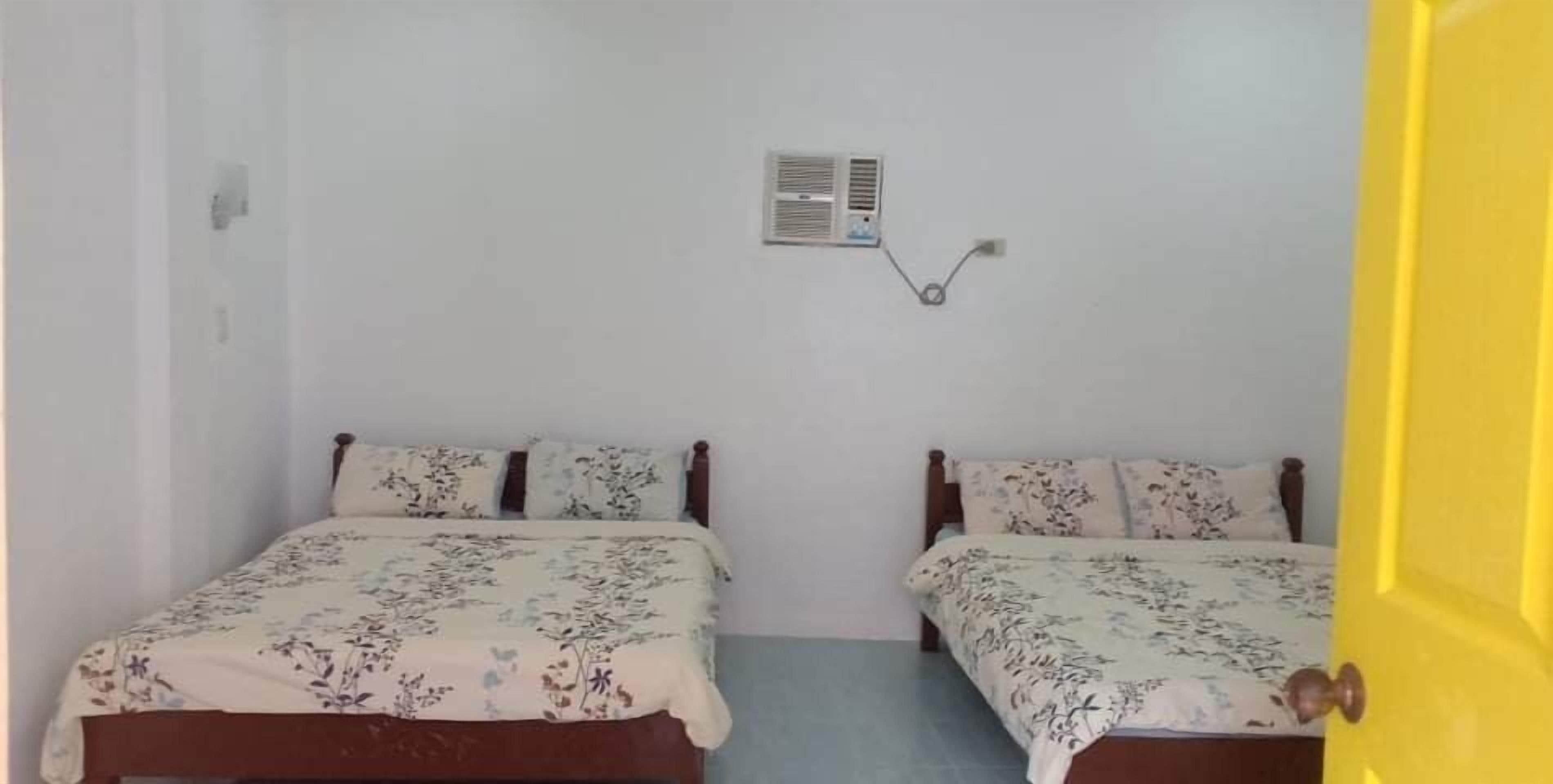 Une chambre avec deux lits en bois, chacun agrémenté d’une literie à motifs floraux, un petit climatiseur fixé au mur et une porte jaune.