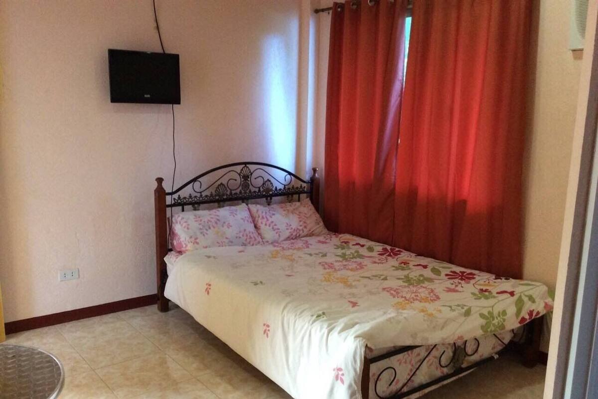 Quarto com uma cama, televisão fixada na parede e janela com cortinas vermelhas.