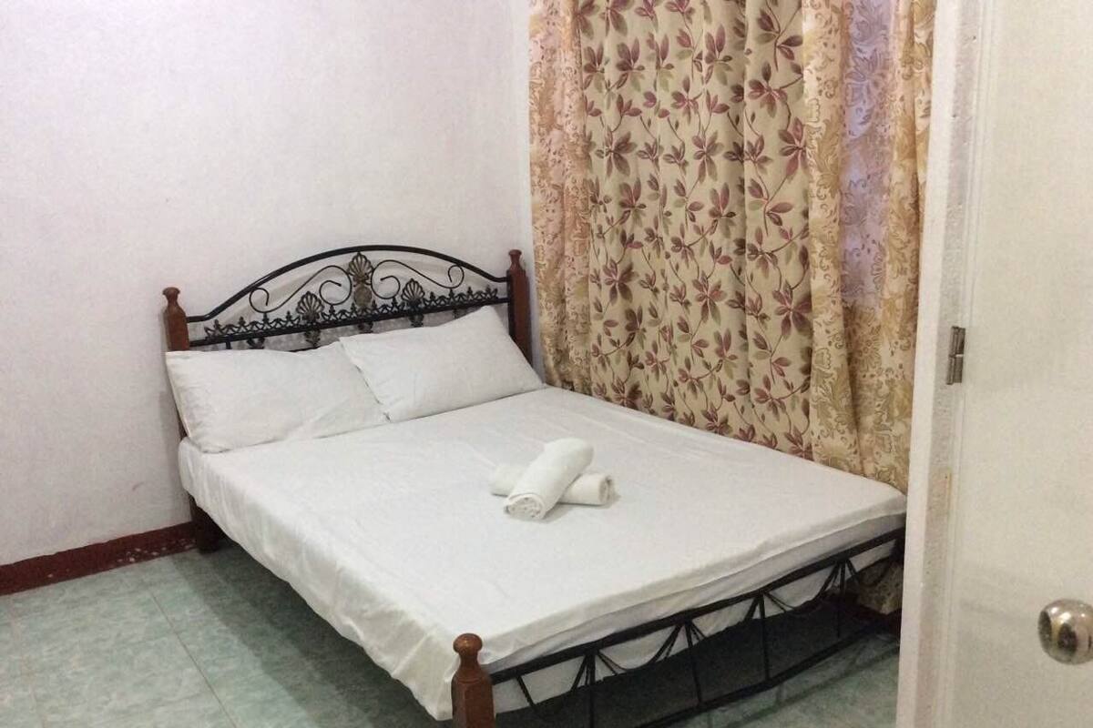 Uma cama de solteiro com estrutura de metal, roupa de cama branca e uma toalha enrolada sobre a cama.
