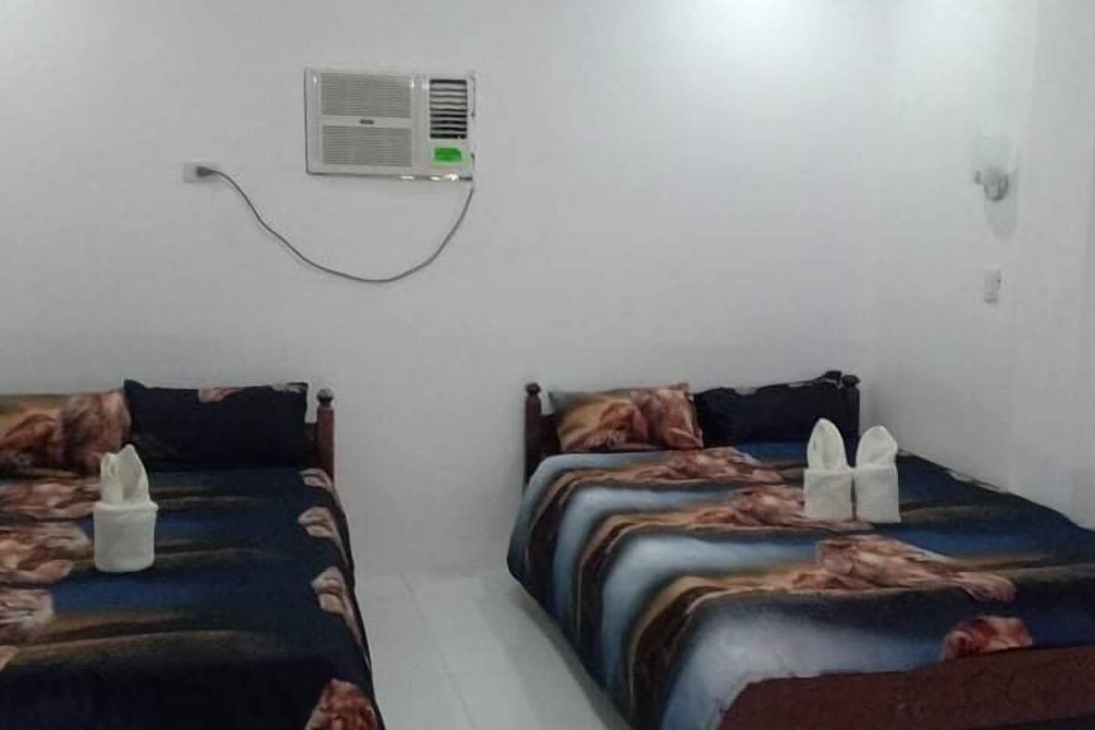 Quarto com duas camas, cada uma com colcha e almofadas estampadas. Há um aparelho de ar condicionado fixado na parede e uma tomada.