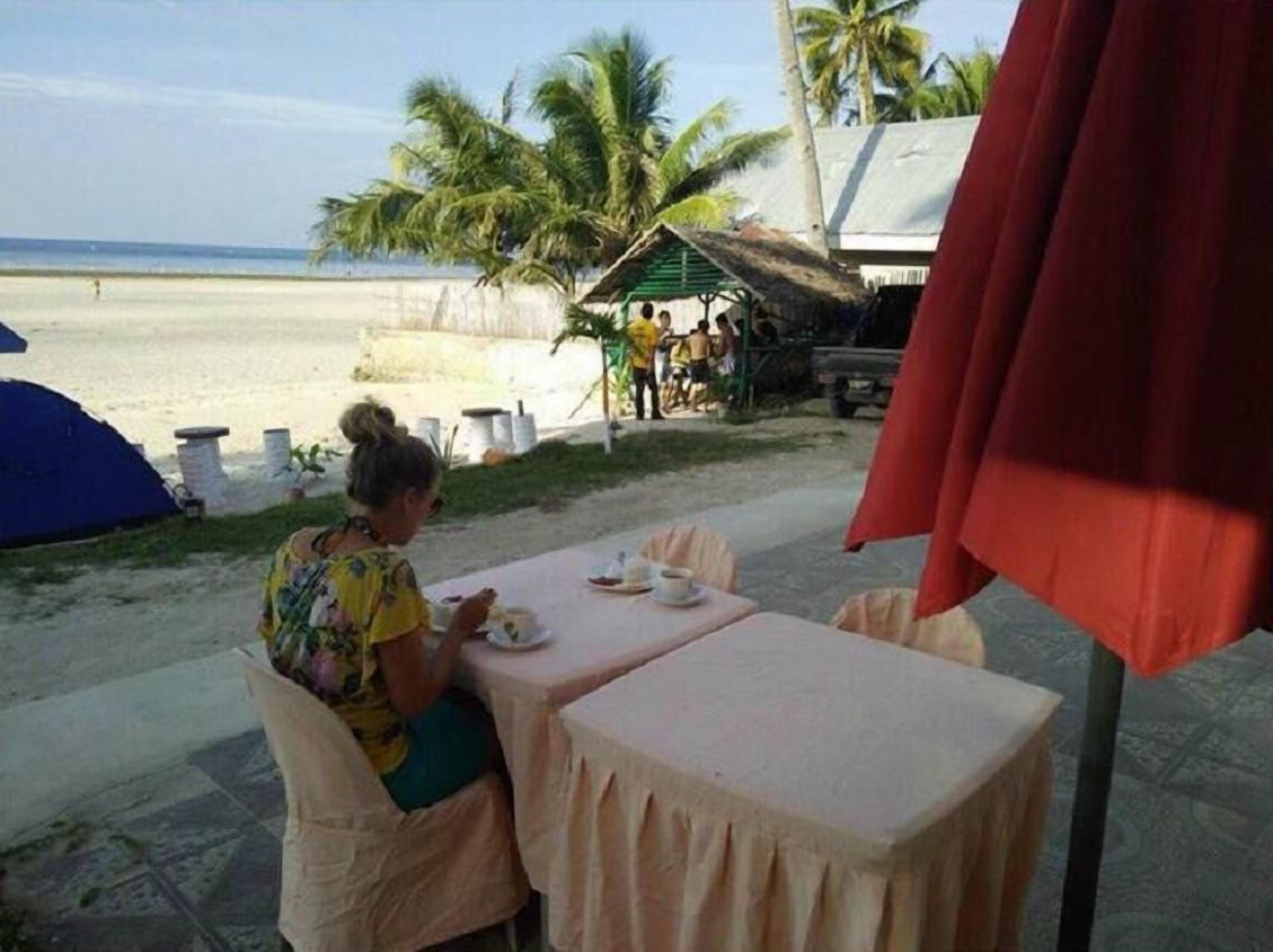 A beachside dining area with a table and chairs, a tent, and a small structure with people in the background.