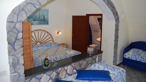 A room with a stone archway, a bed with a blue and white patterned bedspread, a small table with a bottle and glasses, and a blue sofa.