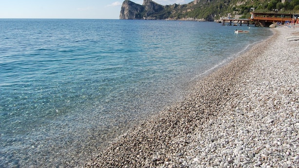 A pebbled beach with clear water, rocky cliffs, and a wooden pier.