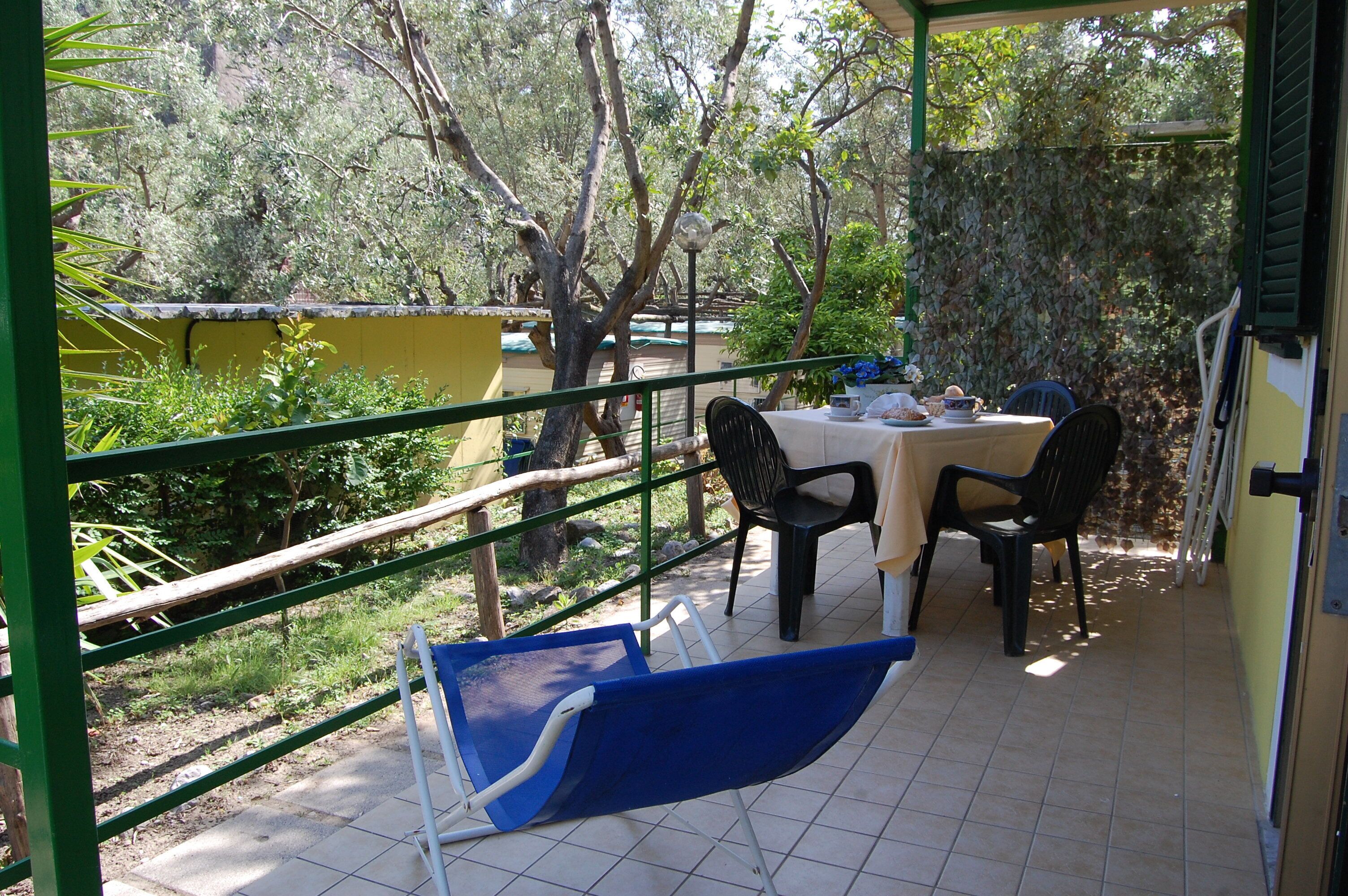 Une terrasse extérieure couverte, avec une table et des chaises, entourée de verdure et bordée d’un bâtiment jaune.