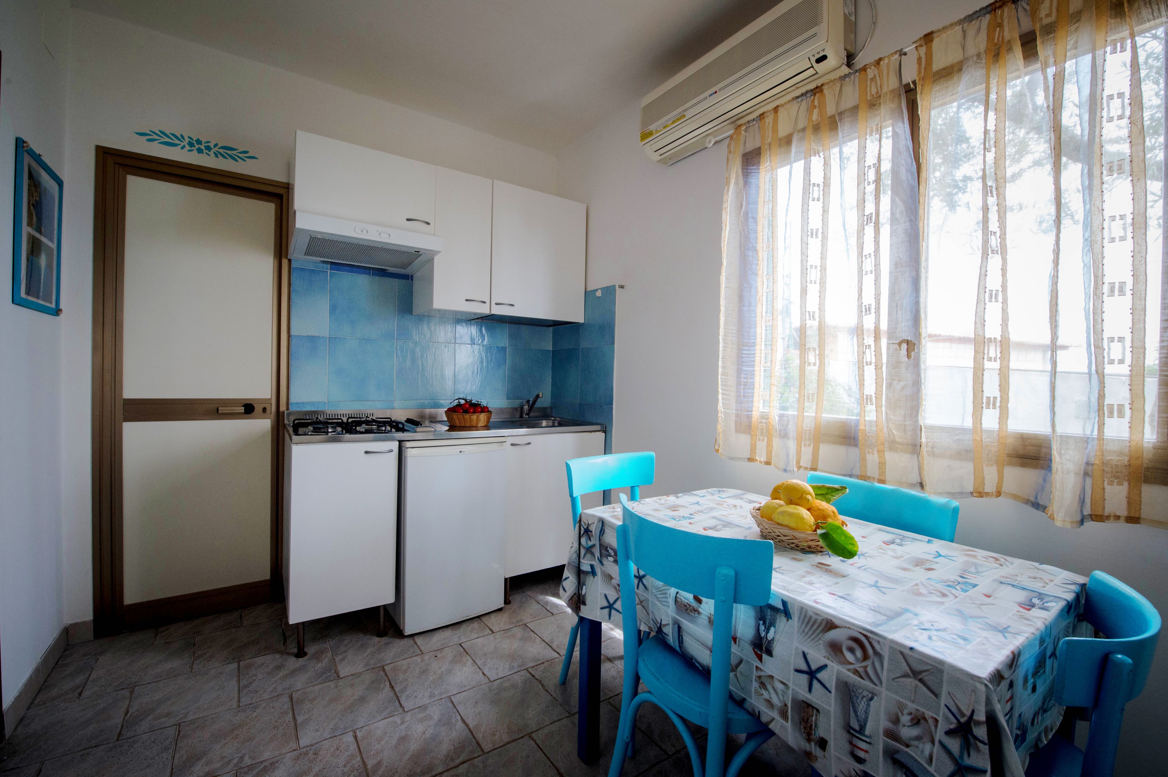A compact kitchen with white cabinets, a gas stove, and a sink. There is a dining table with a blue chair and a tablecloth with a nautical theme.
