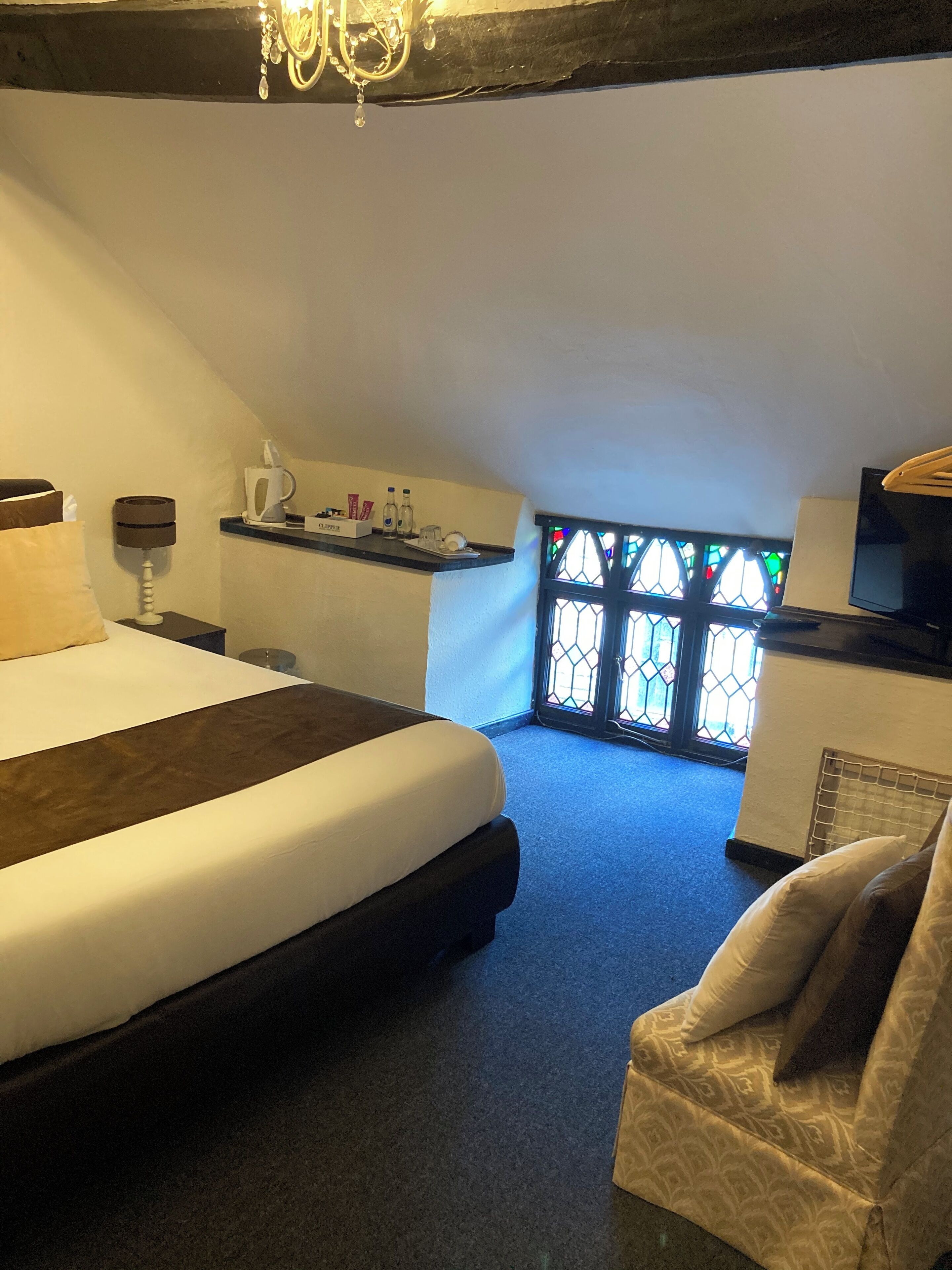 Ein Schlafzimmer mit einem großen Bett, einem kleinen Tisch mit Wasserkocher, einem Sessel und einem Fenster mit Buntglas.