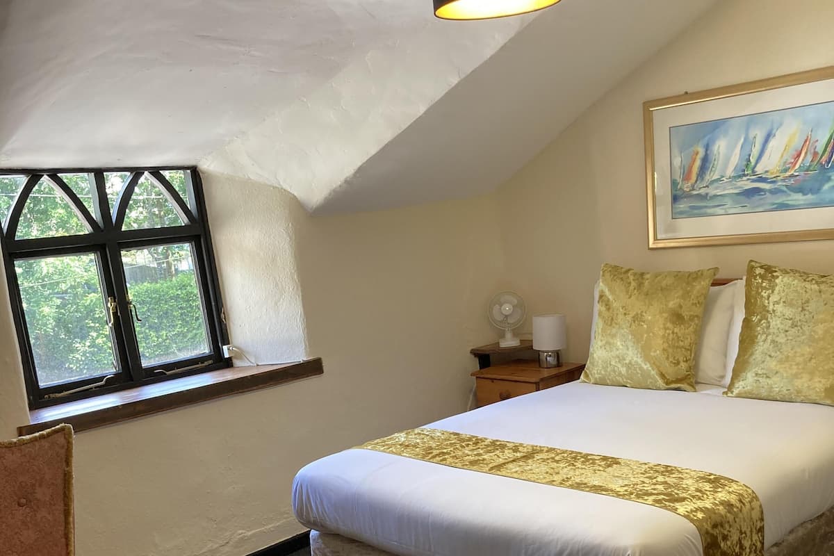 A bedroom with a large bed, a chair, a nightstand, a lamp, a painting, and a window with a view of greenery.
