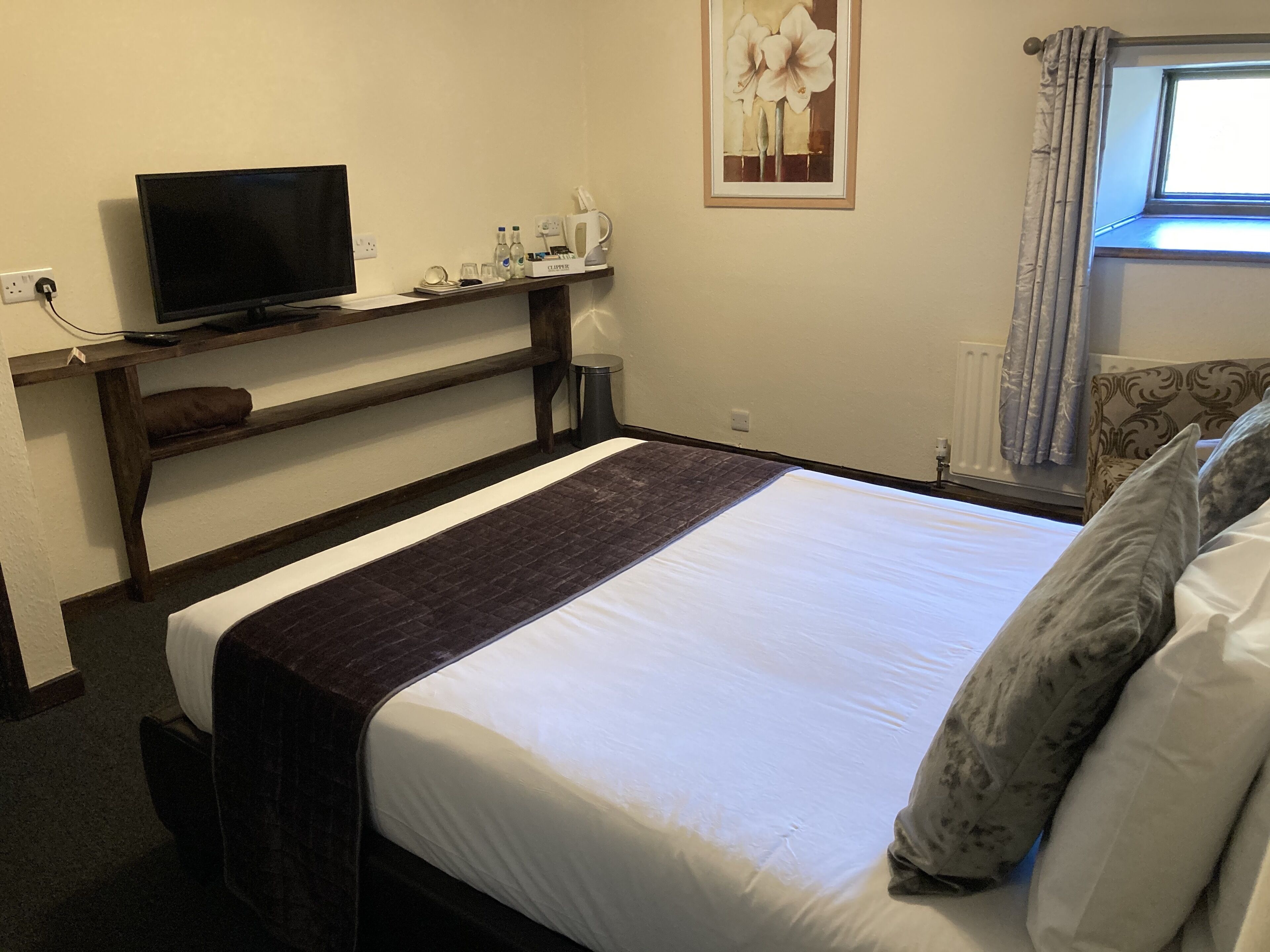 Une chambre d’hôtel avec un lit, un téléviseur posé sur une étagère en bois, une fenêtre avec des rideaux et un tableau accroché au mur.