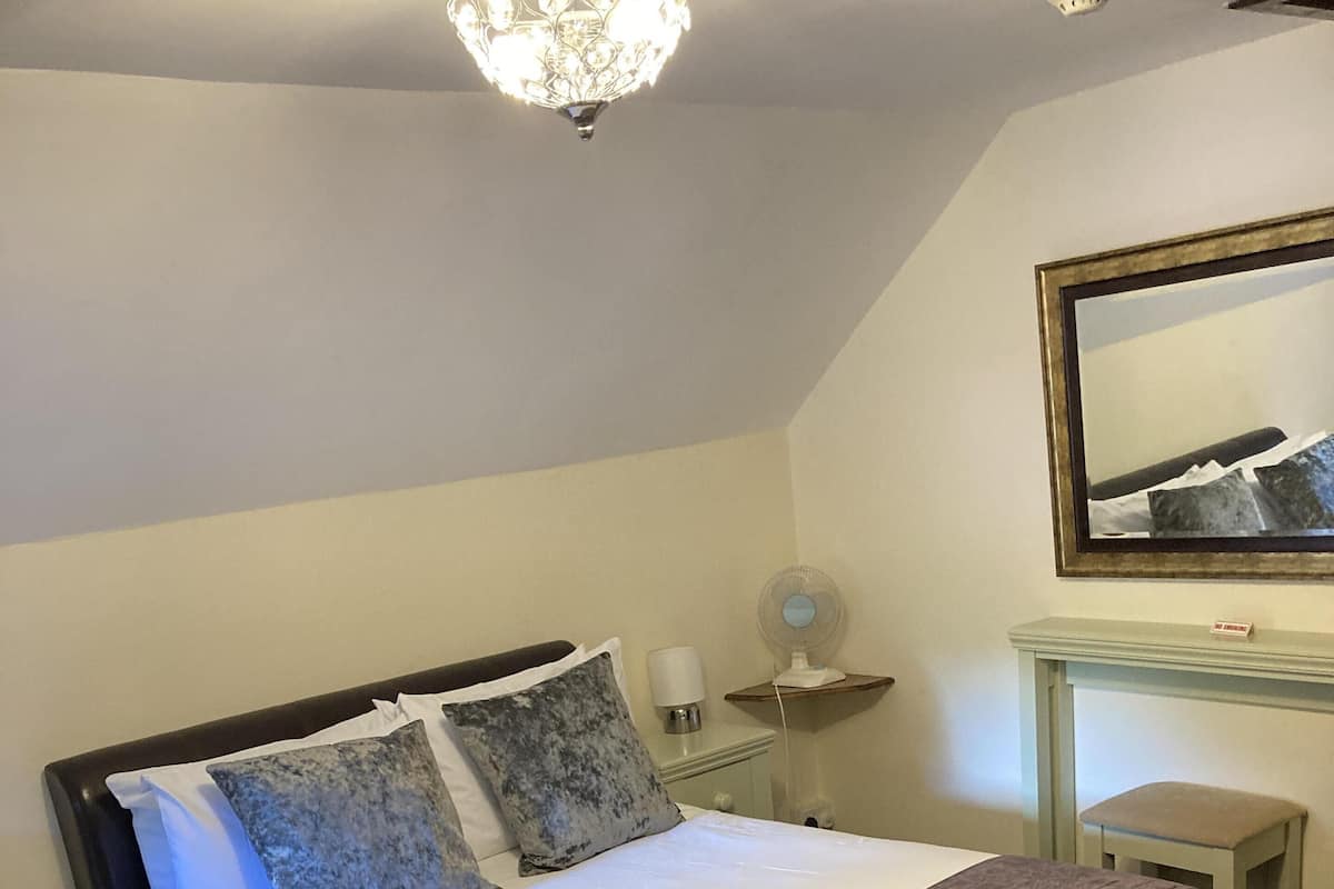 A neatly made bed with a white and purple bedspread, a bedside table with a lamp, and a small stool.