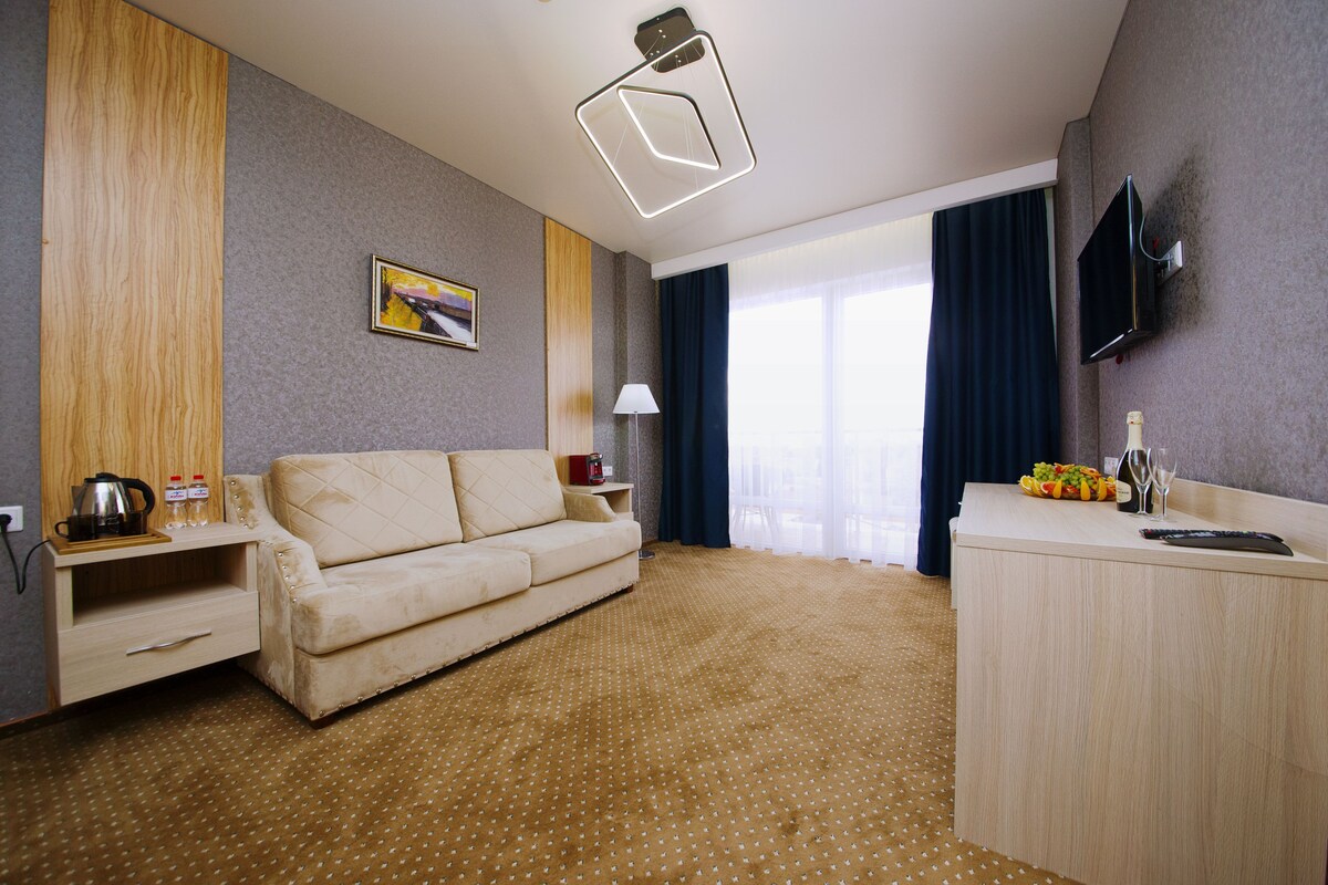 A hotel room with a sofa, a desk, a flat-screen TV, a lamp, and a window with curtains.