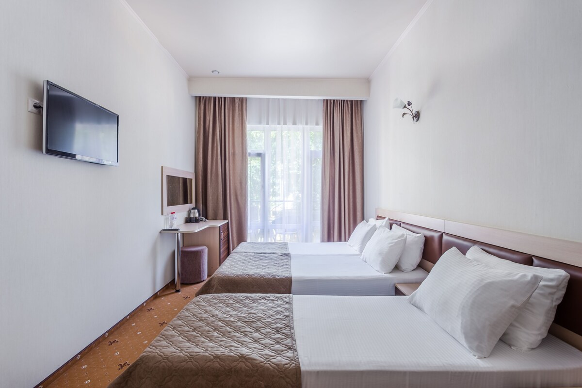 A hotel room with two beds, a TV, a desk, and a window with curtains.