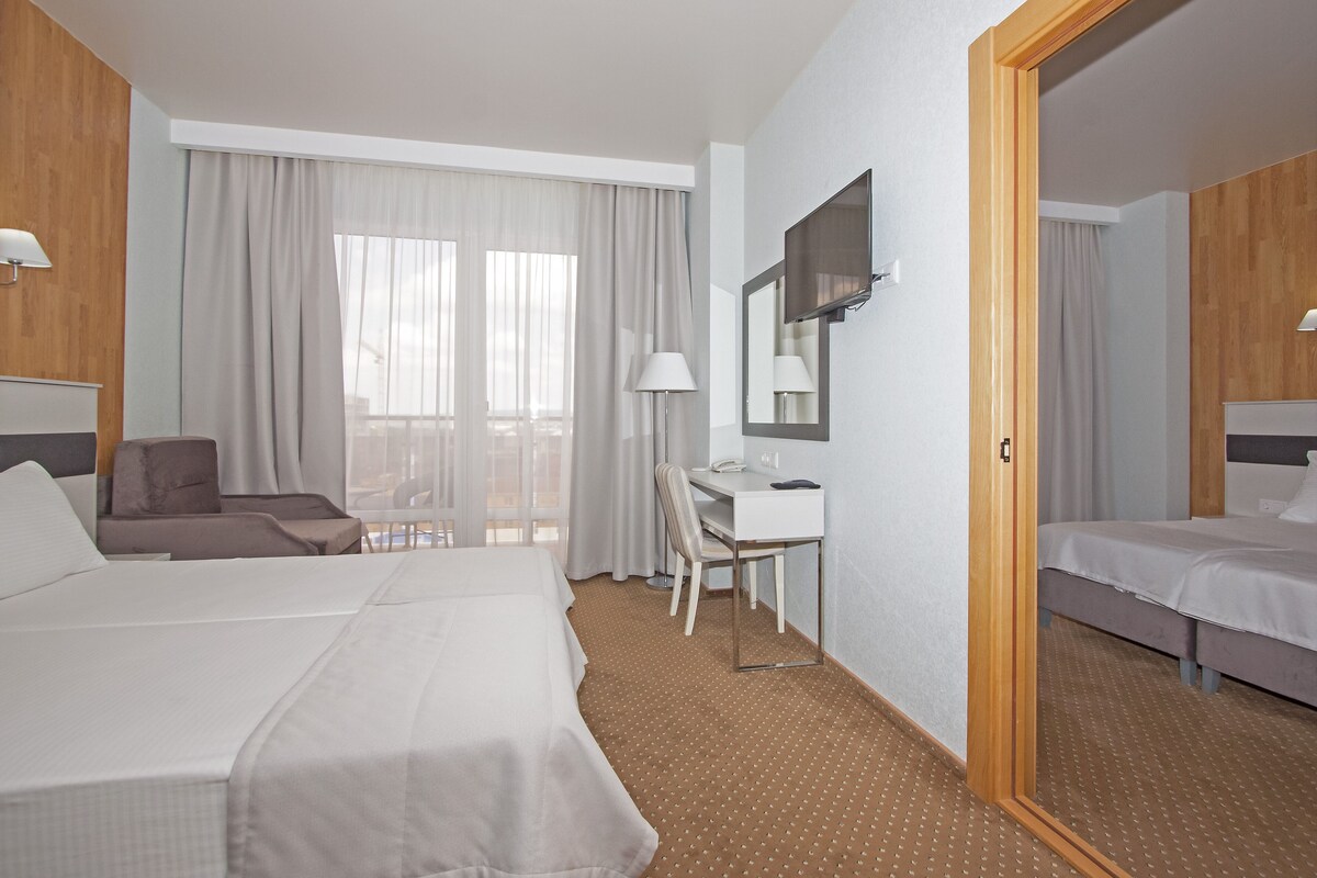 A hotel room with two beds, a desk, a chair, and a large window with curtains.