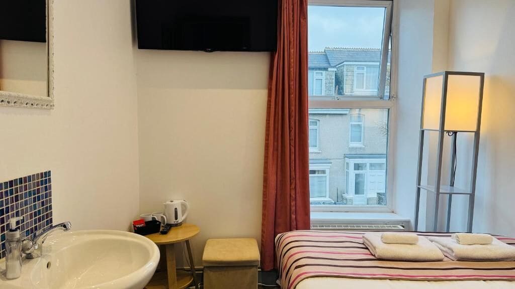 A hotel room with a bed, a bedside table with a kettle, a sink, a mirror, a window with curtains, and a TV mounted on the wall.
