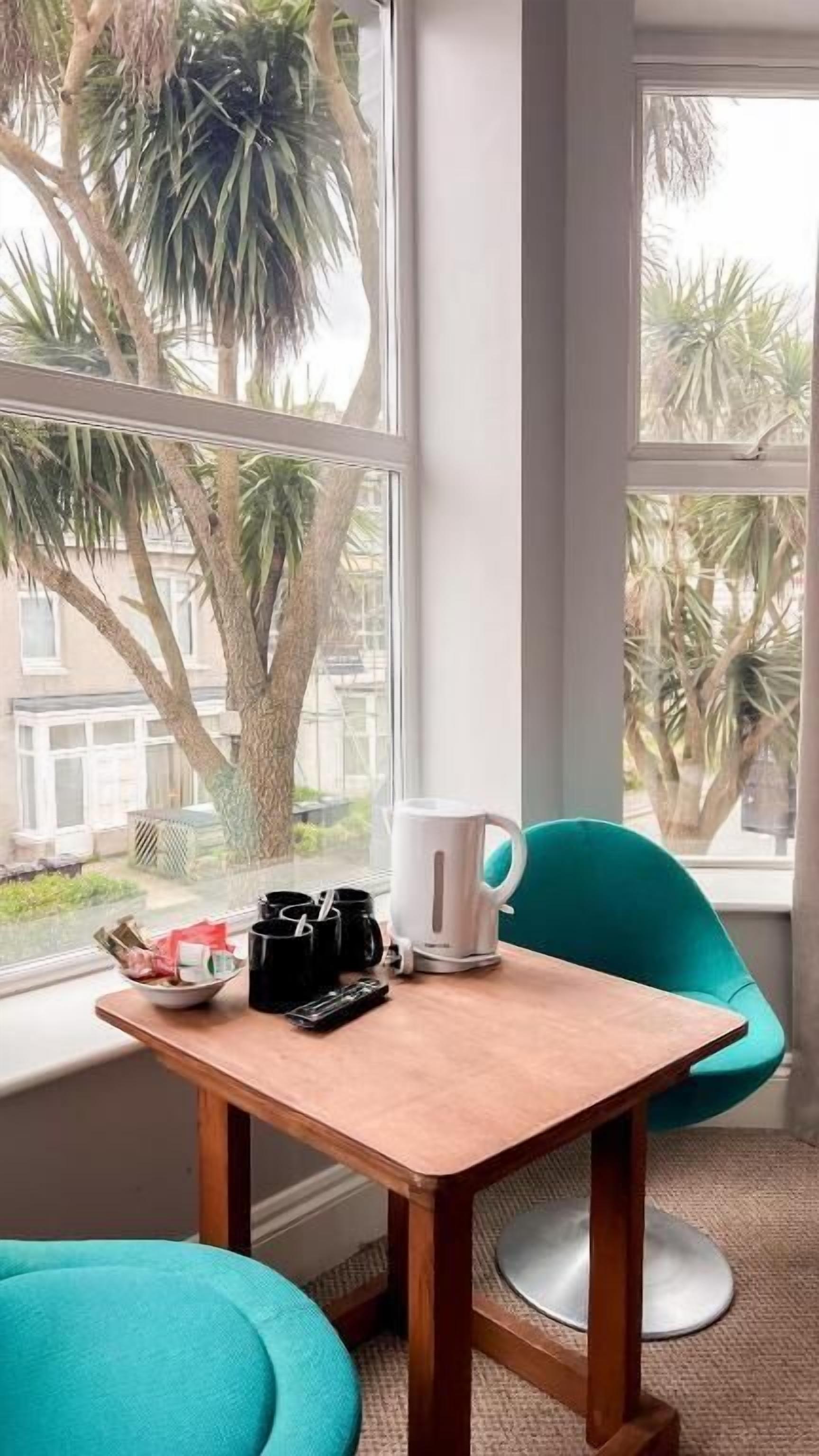 A room with a wooden table, a white kettle, a black mug, and a teal chair by a window with a view of palm trees.