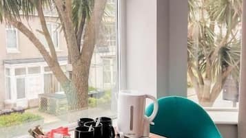 A room with a wooden table, a white kettle, a black mug, and a teal chair by a window with a view of palm trees.