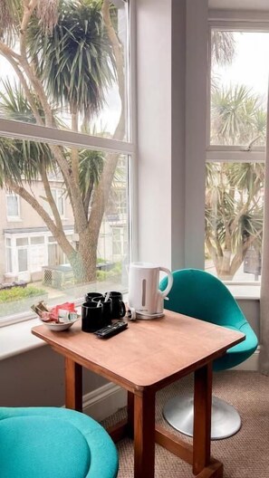 Quarto com uma mesa de madeira, uma chaleira branca, uma caneca preta e uma cadeira azul-turquesa perto de uma janela com vista para palmeiras.