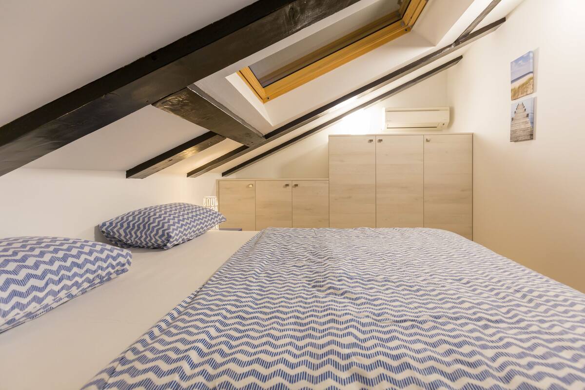 Un dormitorio con techo inclinado, vigas de madera, cama con ropa de cama estampada, un armario y un tragaluz.